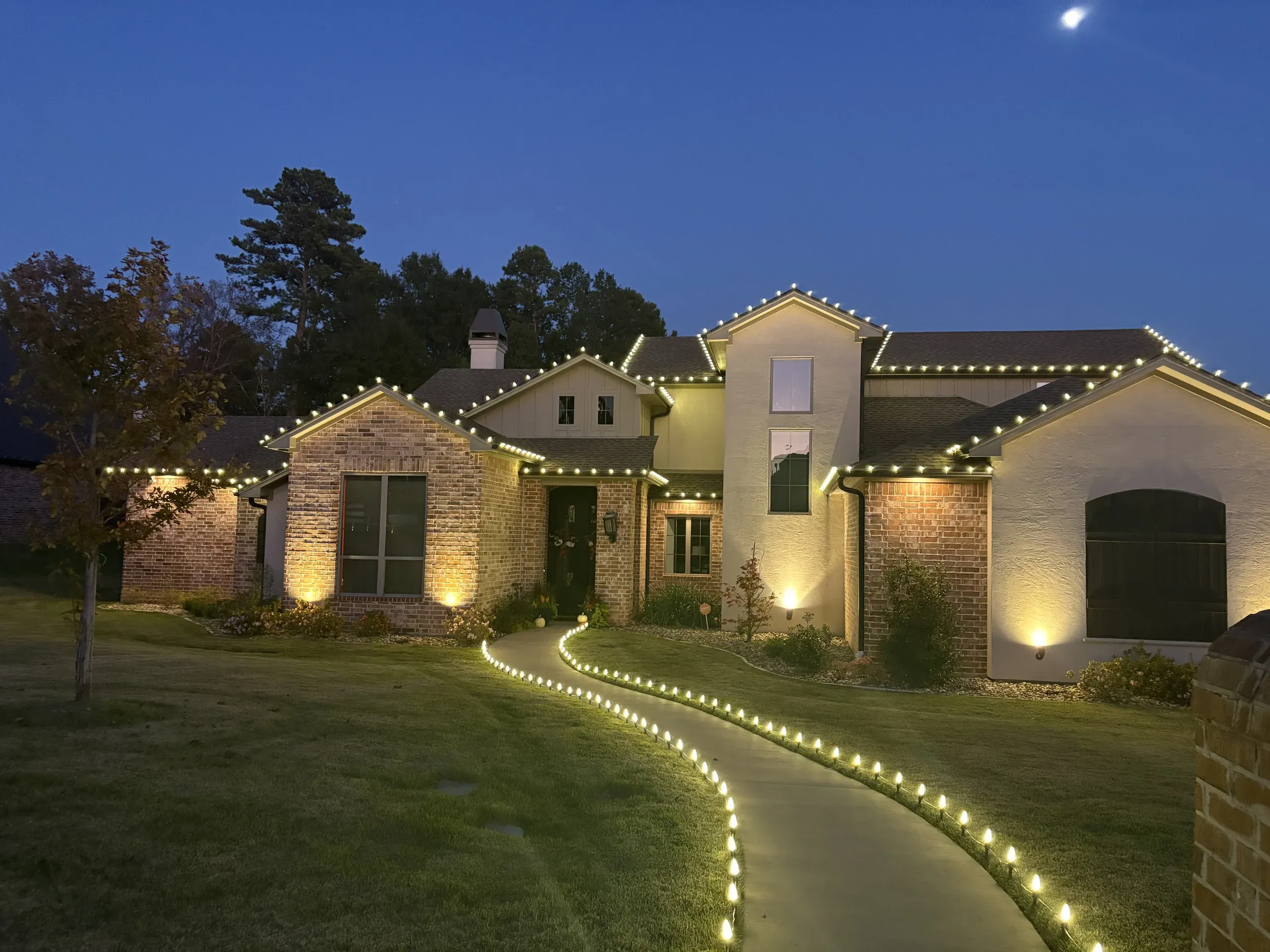 A brick and stucco home in Dallas-Fort Worth with warm white C9 lights along every roofline and a lit front walkway at dusk