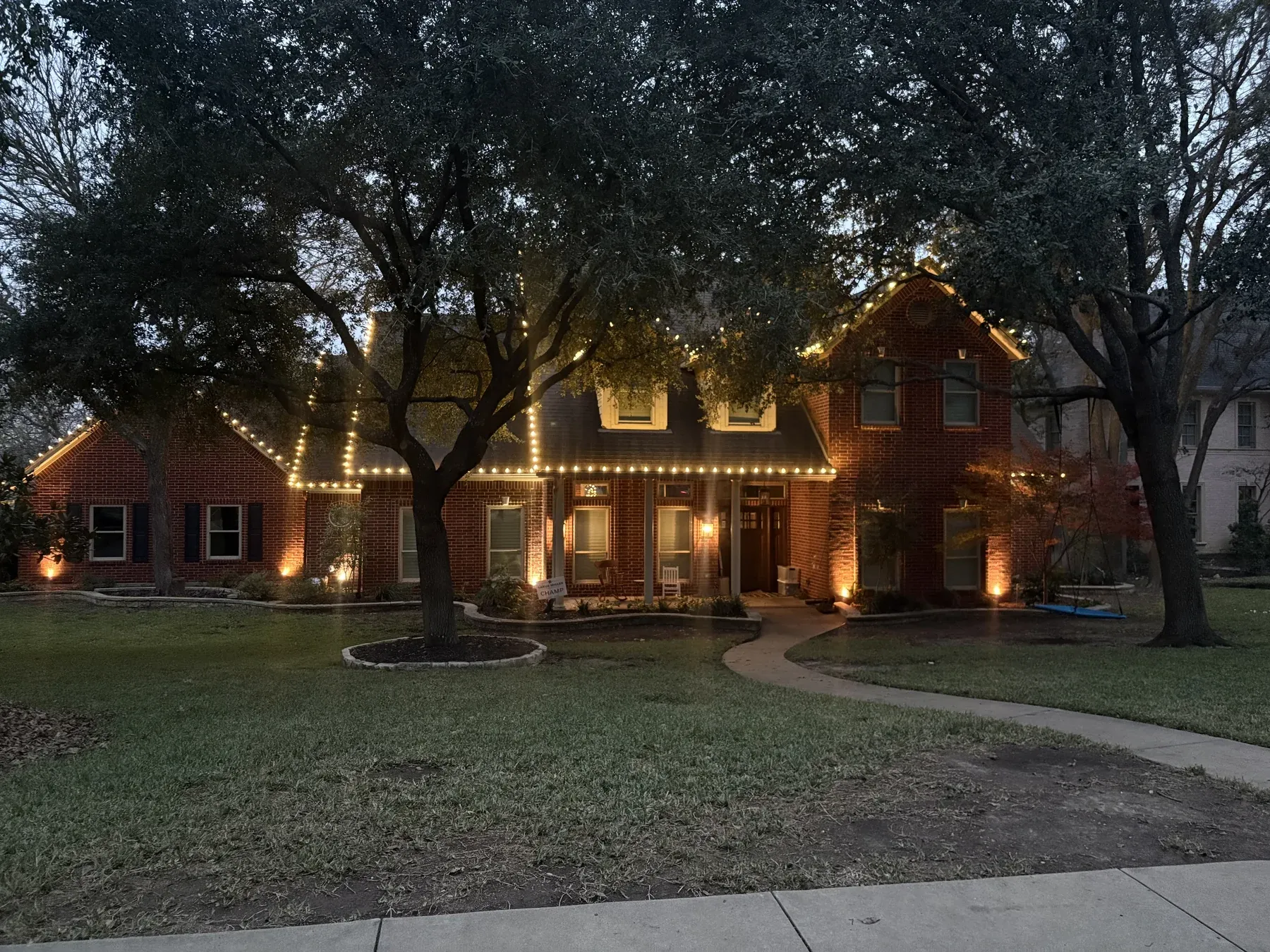Warm white roofline lights on a two story red brick home framed by oak trees