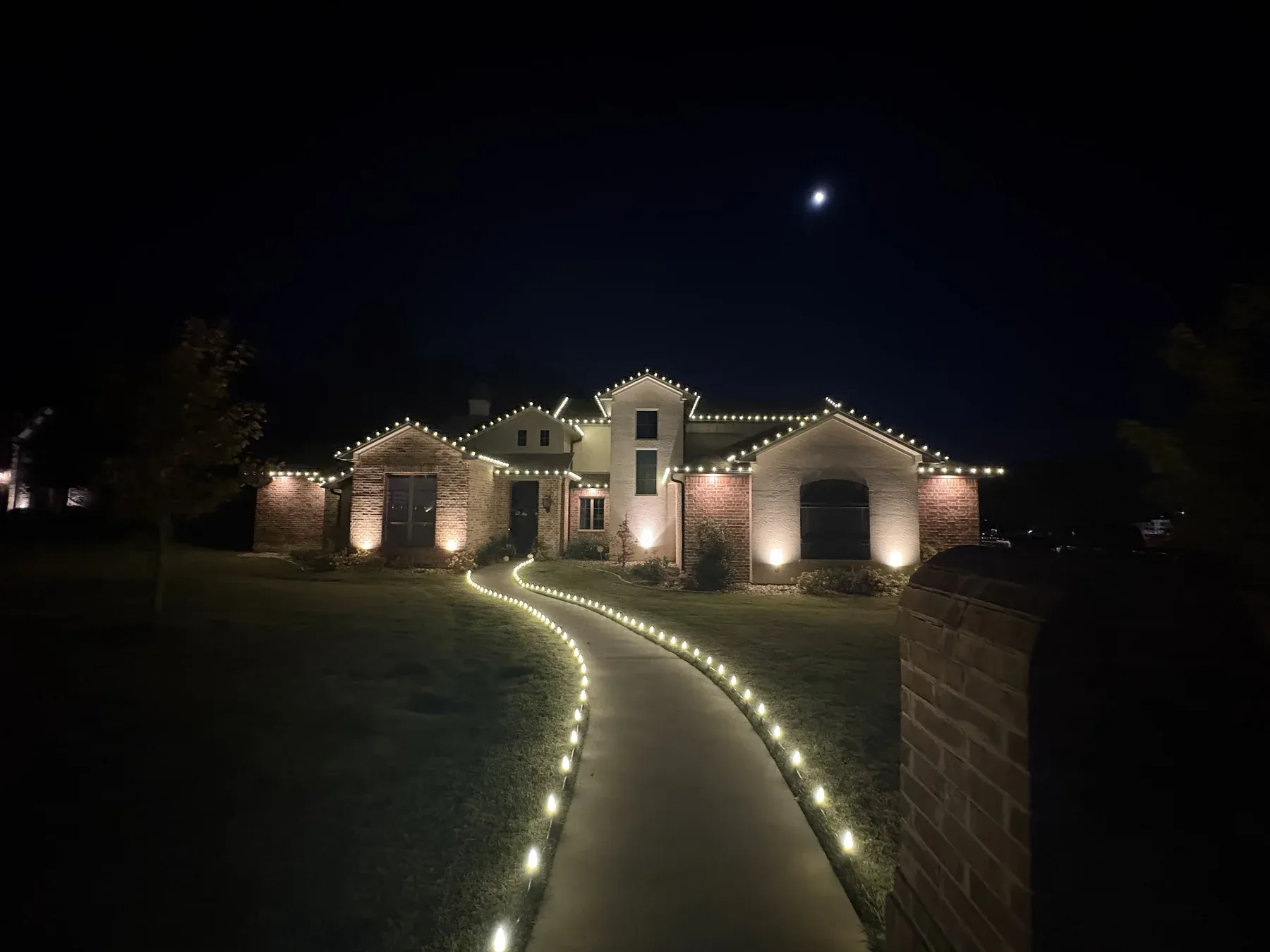 Christmas lights outlining the roof peaks and front walkway of a single story brick home at night